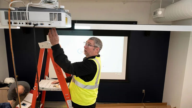 AV support, maintenance and upgrades Technician on a ladder servicing a ceiling-mounted projector in a meeting room