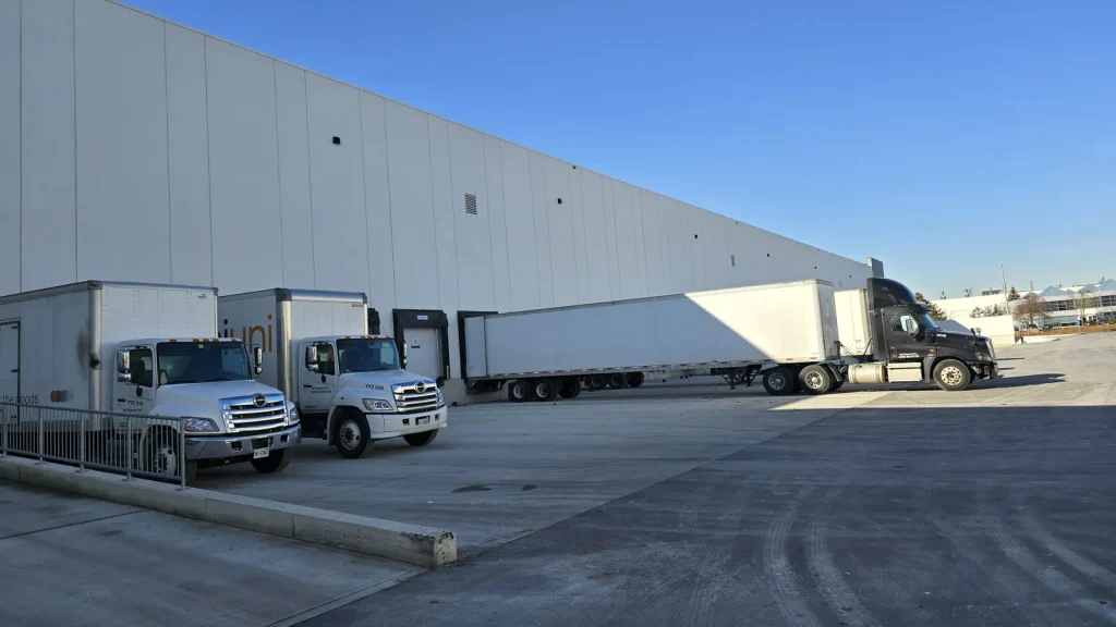Box trucks and a tractor-trailer backed into dock doors along a large warehouse wall